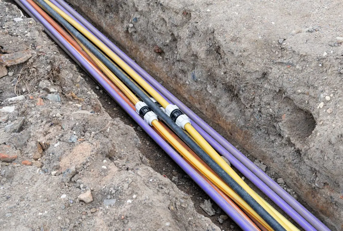 Several colored utility pipes, including yellow, purple, and black, are laid in a trench in the ground, likely for electrical or communication infrastructure.
