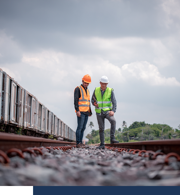 Two workers wearing safety vests and helmets inspect railroad tracks outdoors, with train cars visible on the left and cloudy sky overhead.